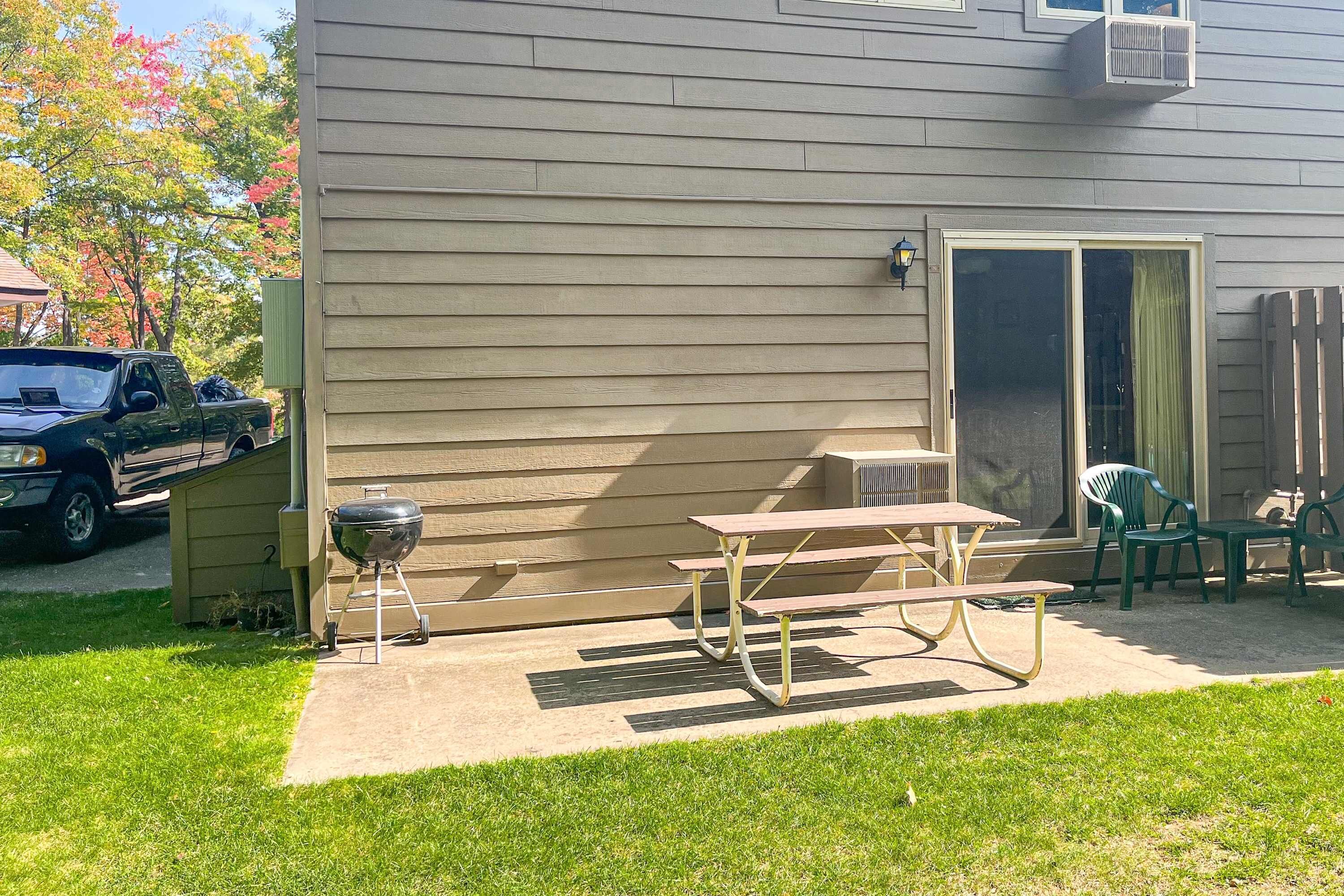 A patio with a grill, a table, and chairs, a sliding glass door, and a vehicle parked in the background.