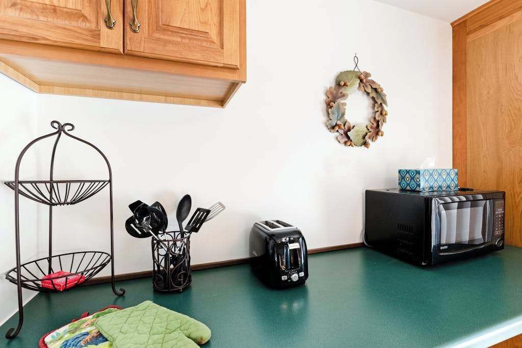 A kitchen with wooden cabinets, a green countertop, a toaster, a microwave, a metal rack with utensils, and a decorative wreath on the wall.