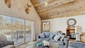 Een ruime woonkamer met een groot raam, houten plafond, een zithoek, een salontafel en een boekenkast.