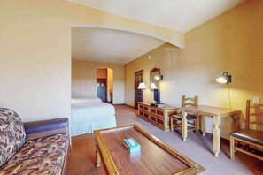 A hotel room with a bed, a sofa, a wooden coffee table, a desk with chairs, and a television.