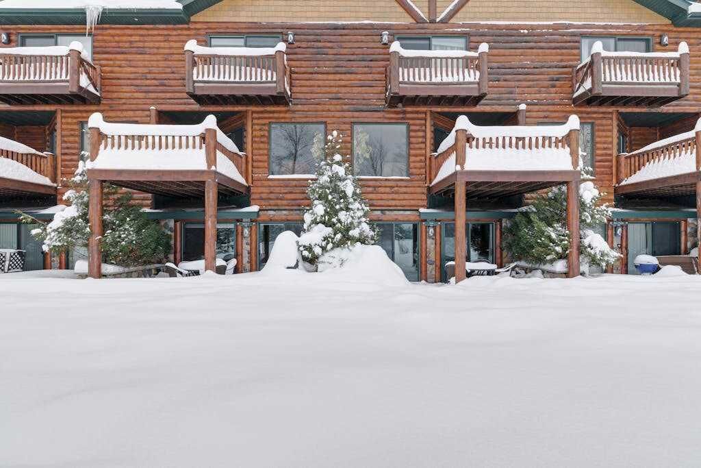 Un chalet en bois à plusieurs étages, doté de balcons, avec un arbre central recouvert de neige et un sol également enneigé.