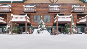 Una cabaña de madera de varios pisos con balcones, un árbol cubierto de nieve en el centro y el suelo nevado.