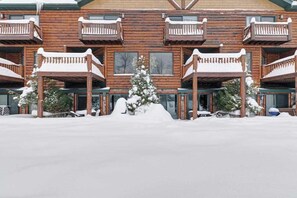 A multi-story wooden lodge with balconies, a central snow-covered tree, and a snow-covered ground.
