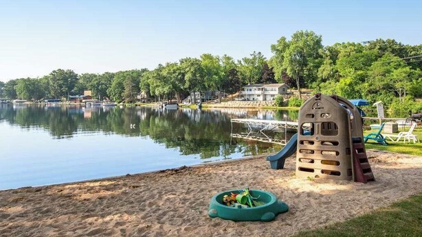 A lakeside park with a playground, a sandy area, and a house in the background. - Inviting lakefront stand-alone condo with private beach, boating, & playground (Wisconsin Dells)