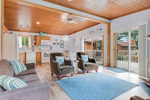 A spacious living area with a wooden ceiling, a kitchen in the background, and a view of a deck.