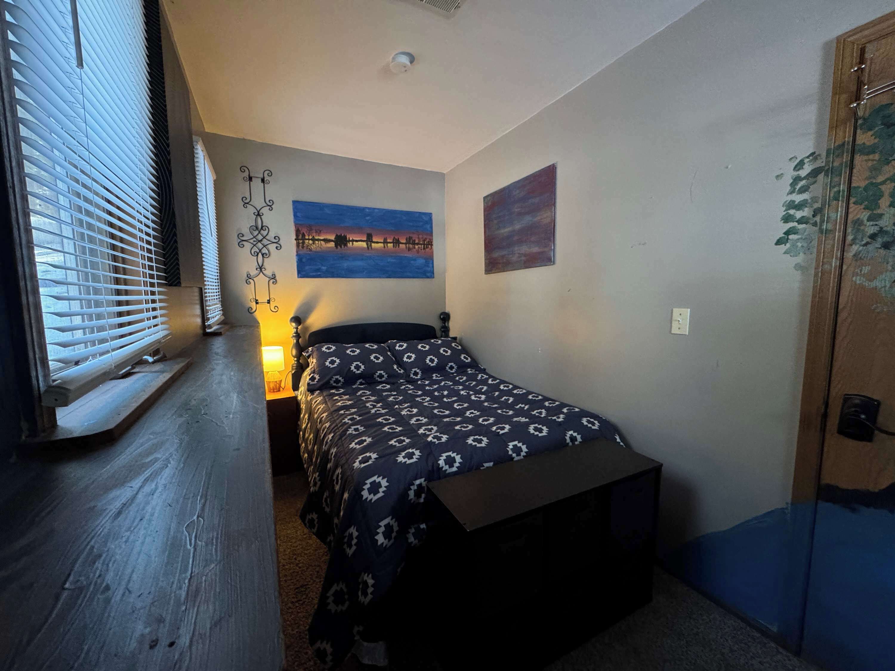 Un dormitorio con cama, mesita de noche, lámpara de pared y dos cuadros.