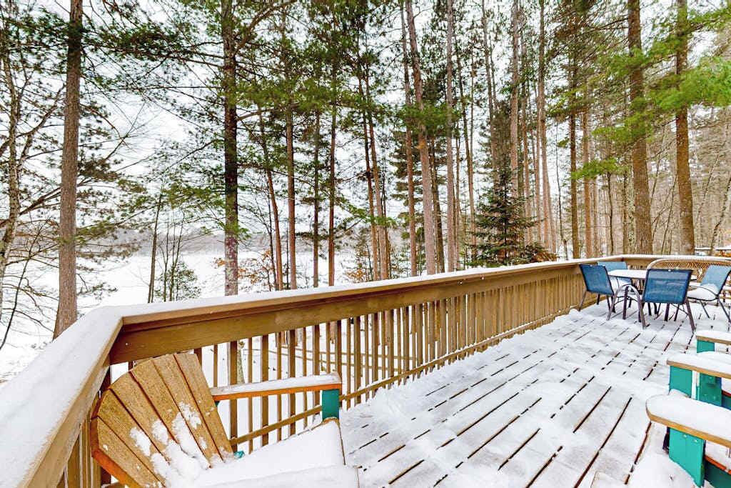 A wooden deck with chairs and tables, surrounded by tall trees and snow.