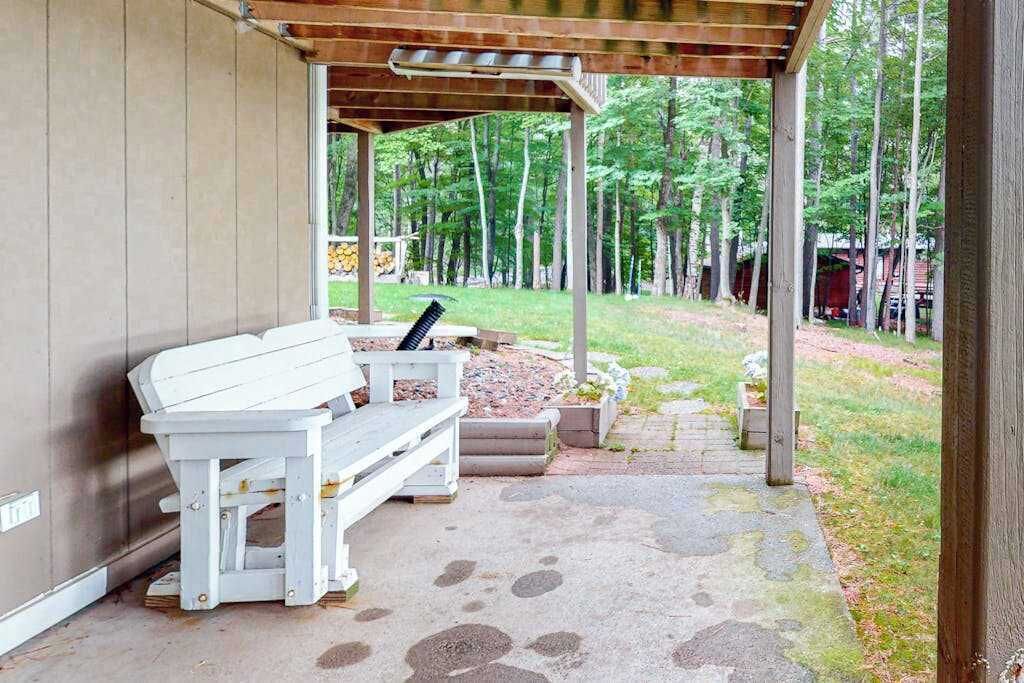 A covered outdoor seating area with a bench and a table, surrounded by a stone pathway and a forested area.