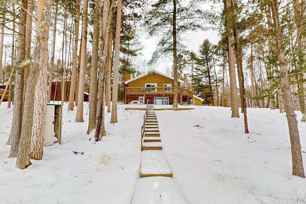 A lodge with a staircase leading to a wooden deck, surrounded by tall pine trees and snow-covered ground.