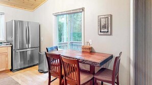 Uma cozinha com geladeira de aço inoxidável, mesa de jantar de madeira e cadeiras, janela com vista para árvores e um quadro na parede.