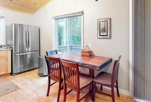 A kitchen with a stainless steel refrigerator, wooden dining table, and chairs, a window with a view of trees, and a framed picture on the wall. - Hillis House | Lakefront house in Arbor Vitae w/water toys & two kitchens (Arbor Vitae)