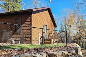 A wooden cabin with a fenced yard, a stone wall, and a clear sky. - Lakefront escape on Sugar Maple w/ dock, firepit, kayaks, & gas grill- Dogs-OK (Phelps)