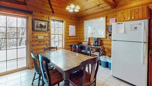 Interno di una baita in legno con un tavolo da pranzo, sedie, un frigorifero e finestre che offrono vista su alberi innevati.