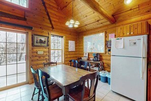 A wooden cabin interior with a dining table, chairs, a refrigerator, and windows with views of snow-covered trees. - Cozy cabin w/ fireplace, whirlpool tub, access to private dock, & fire pit (Mercer)
