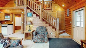 Interno di una baita in legno con una scala, una poltrona a fantasia, un divano e un tavolino con una lampada.