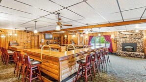 A log cabin interior with a central wooden bar, multiple bar stools, a stone fireplace, and a ceiling fan. - Full hotel with bar, commercial kitchen, pool table & fireplaces - dog-friendly (Winchester)