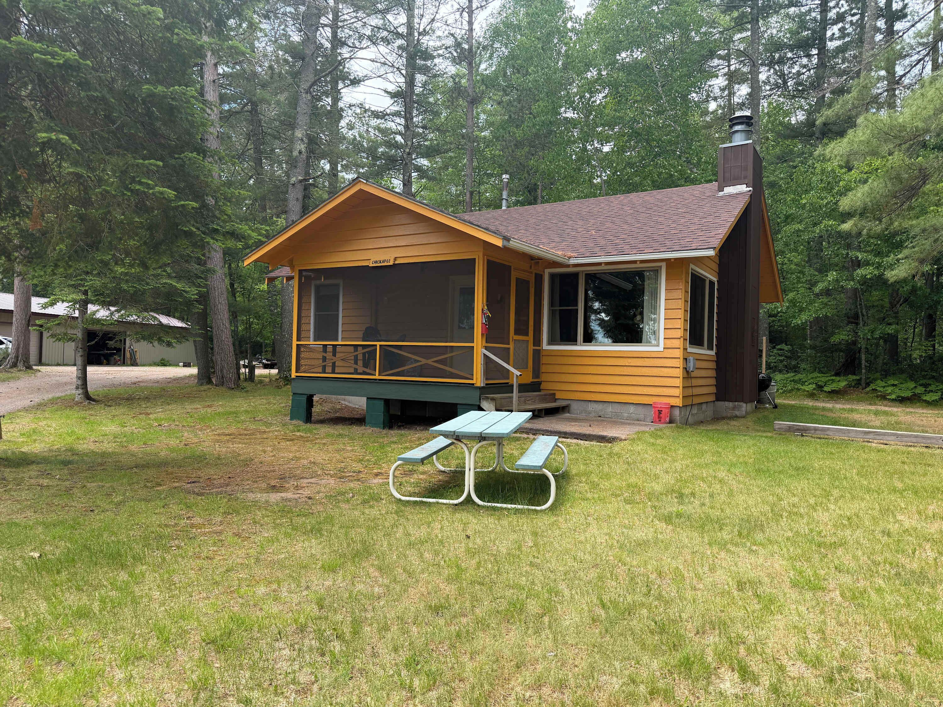 A cabin with a porch, a chimney, and a picnic table in a wooded area.