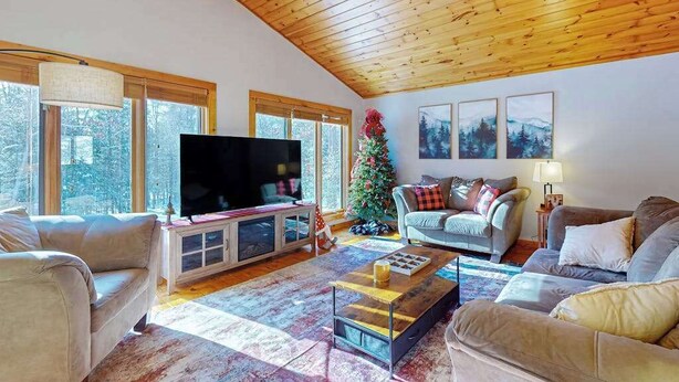 A cozy living room with a wooden ceiling, large windows, a flat-screen TV, and a Christmas tree. - Serene Home w/ large fenced in yard, jetted tub, WIFI, near Lakes & Trails (St. Germain)