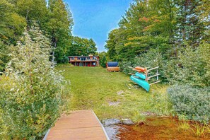 A wooden deck leading to a house surrounded by dense greenery. - Dog-friendly lakefront home with boat access, deck, fireplace, gas grill, kayaks (Winchester)