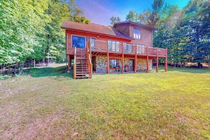 A two-story wooden cabin with a balcony, surrounded by a grassy yard and trees. - Beautiful Lakefront Cabin with a Private Dock, Firepit, and Large Yard! (Presque Isle)