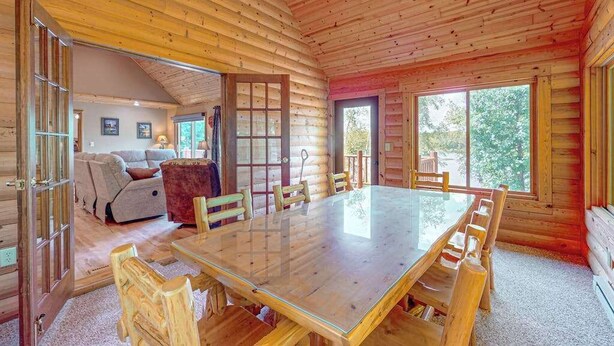 Interior de uma cabana de toras com uma mesa de jantar, cadeiras, um sofá e uma janela grande.