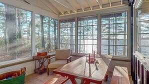 Un espace extérieur aménagé, abrité et doté d’une table et de bancs en bois, de grandes fenêtres et d’une vue sur les arbres et l’eau.