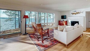 Un salon spacieux avec un canapé blanc, un fauteuil à bascule, une cheminée et de grandes fenêtres donnant sur un plan d’eau.