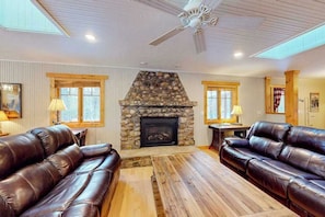 A cozy living room with a stone fireplace, leather sofas, wooden flooring, and a ceiling fan.