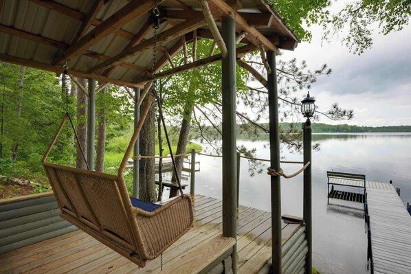 A wooden deck with a swing chair, a bench, and a dock extending into a calm lake.