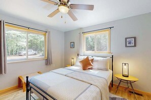 Un dormitorio con una cama, dos ventanas, un ventilador de techo, una mesita de noche y una mesilla.