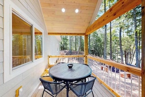 Un espacio exterior techado con una mesa redonda y sillas, ventanas amplias y vistas a un bosque.