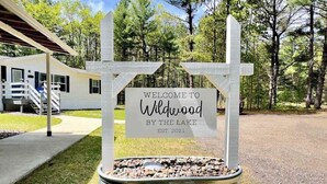 A welcome sign in a rustic metal container with a message 'WELCOME TO Wildwood By The Lake EST. 2021' and a white picket fence. - Peaceful 3BR home w/ 3 season clubhouse 1/2 mile from Castle Rock Lake (Mauston)