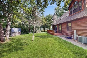 A log cabin with a stone chimney, a covered deck, and a well-maintained lawn. - Waterfront Log Cabin Retreat with Private Boat Dock, Gas Grill, Fire pit & WIFI (Necedah)