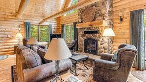 Interior de una cabaña de madera con chimenea de piedra, muebles de madera y ventanas amplias.