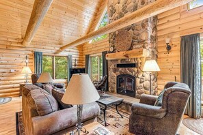 A log cabin interior with a stone fireplace, wooden furniture, and large windows.