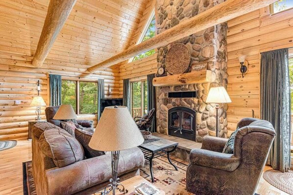 A log cabin interior with a stone fireplace, wooden furniture, and large windows.