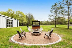 A circular outdoor seating area with a fire pit, surrounded by Adirondack chairs, in a grassy yard with trees and a building in the background. - Renovated barn retreat with firepit, grill, sauna, foosball, kayaks & movie room (Necedah)
