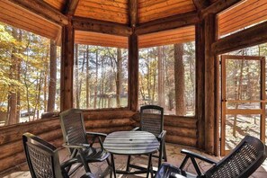 A wooden cabin with a round table and chairs, large windows, and a view of a forest.