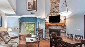 A living room with a fireplace, a television mounted on the stone wall, a dining table with chairs, and a sofa. - Cozy Fireside Condo with Jetted Tub & AC (Wisconsin Dells)