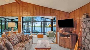 A cozy living room with a large window, wooden walls, a television, and a stone fireplace. - Lakefront home w/ deck, grill, fireplace, firepit, AC, & WiFi (Eagle River)