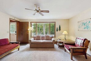A cozy living room with a ceiling fan, a sofa, a chair, a small table, and a window with a view of trees. - Lovely, family-friendly home w/ a patio, balcony, forest views, & river access (Eagle River)