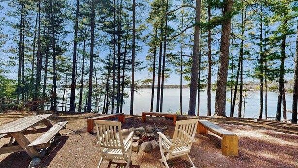 Ein Campingplatz mit Feuerstelle, Holzsesseln und Picknicktischen am See, umgeben von hohen Bäumen.