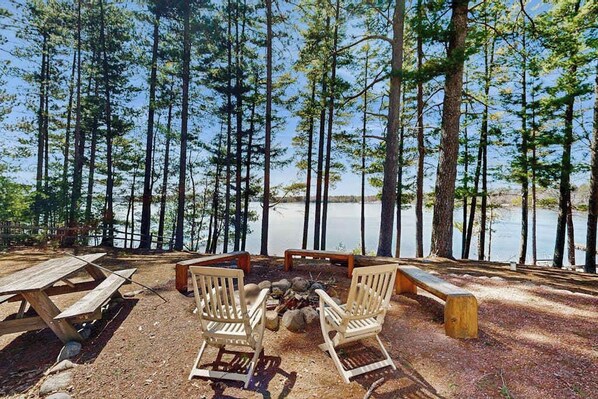 Un camping con una fogata, sillas de madera y mesas de picnic junto a un lago rodeado de árboles altos.