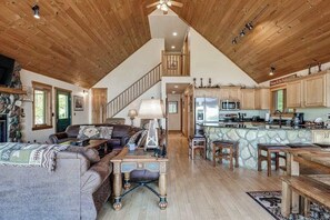 A spacious loft-style living area with a high ceiling, wooden beams, a stone fireplace, a dining area with a stone island, and a kitchen with modern appliances. - Lakefront lodge with private dock, firepit, deck & sunset views (Eagle River)