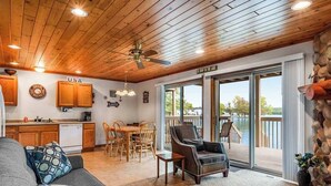 A cozy living area with a wooden ceiling, a sofa, a dining table with chairs, a kitchen with wooden cabinets, and a view of a lake through glass doors.