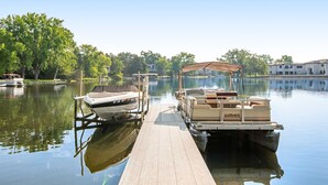 A dock with a pontoon boat and a motorboat, calm water, and trees in the background.