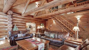 Interior de uma cabana de tronco com escada de madeira, sofás de couro, mesa de jantar e cozinha.
