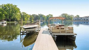 Un quai avec des bateaux, une jetée en bois et un lac paisible, avec des arbres et des bâtiments en arrière-plan.