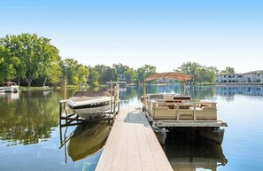 A dock with boats, a wooden pier, and a calm lake with trees and buildings in the background. - Inviting lakefront condo - amazing views, beach access, fireplace, dock, AC, W/D (Wisconsin Dells)