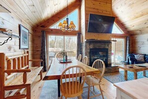 A wooden cabin interior with a dining area, fireplace, and a TV mounted on the wall. - Charming Log Cabin-  Boat, fish, hike & snowmobile—minutes to lakes & trails (Friendship)